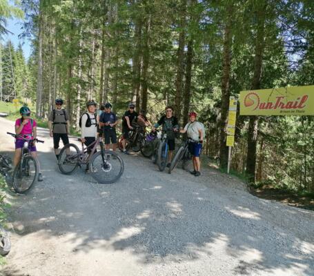 Eine Gruppe Mountainbiker pausiert auf einem sonnigen Waldweg neben einem Trail-Schild. Die Szene zeigt Fahrräder, Sportausrüstung und hohe Nadelbäume – ideal für Outdoor-Fitness, geführte Bike-Touren und aktive Naturerlebnisse.