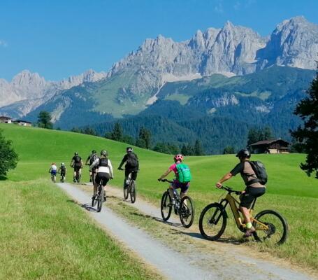 Mountainbiken in den Alpen | © © Bikeacademy Eine Gruppe Mountainbiker fährt auf einem schmalen Schotterweg durch grüne Almwiesen, umgeben von beeindruckenden Alpenbergen und klarem Himmel. Die Szene vermittelt aktive Erholung, Naturgenuss und sportliches Radfahren in malerischer Berglandschaft.