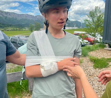 Erste Hilfe nach Radsturz | © © Bikeacademy Ein junger Radfahrer mit Helm steht im Freien vor Bergkulisse, während sein verletzter Arm mit einer provisorischen Schlinge versorgt wird. Die Szene zeigt Erste Hilfe nach einem möglichen Fahrradunfall im sportlichen Outdoor-Umfeld.