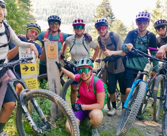 Eine fröhliche Gruppe von Mountainbikern posiert mit Fahrrädern auf einem sonnigen Waldweg. Helme, Rucksäcke und Sportkleidung zeigen die Vorbereitung auf eine gemeinsame Bike-Tour in natürlicher Umgebung.