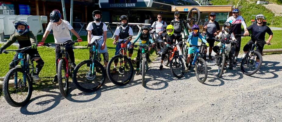 Eine Gruppe junger Mountainbikerinnen und Mountainbiker steht mit Fahrrädern und Helmen vor einer Bergbahnstation. Die sonnige Szene zeigt ein sportliches Outdoor-Erlebnis in alpiner Umgebung, ideal für Bike-Camps, Fahrtechniktraining und gemeinsame Abenteuer am Berg. Turracher Höhe - Kärnten - AUSTRIA