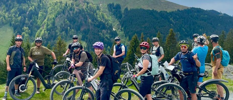 Eine Mountainbike-Gruppe posiert mit Fahrrädern vor grünen Alpenhängen und bewaldeten Bergen. Die sportliche Szene vermittelt Teamgeist, Outdoor-Abenteuer und aktive Erholung in beeindruckender Naturkulisse.